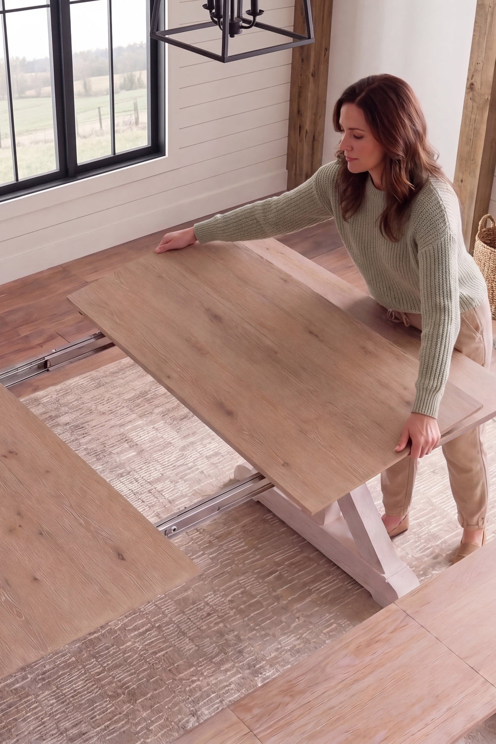 A woman in a sage green sweater and tan pants is shown lifting a belgian oak wood dining table extension panel on a sliding track system in a bright, modern farmhouse dining room.